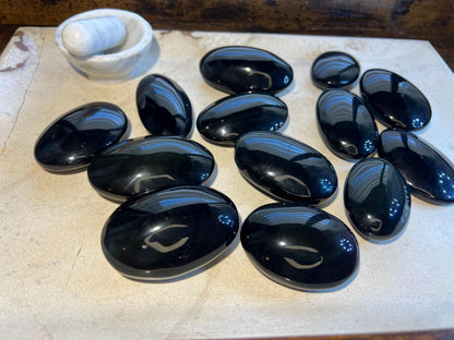 May include: A collection of twelve smooth, black oval-shaped stones. The stones are arranged on a light-colored surface.